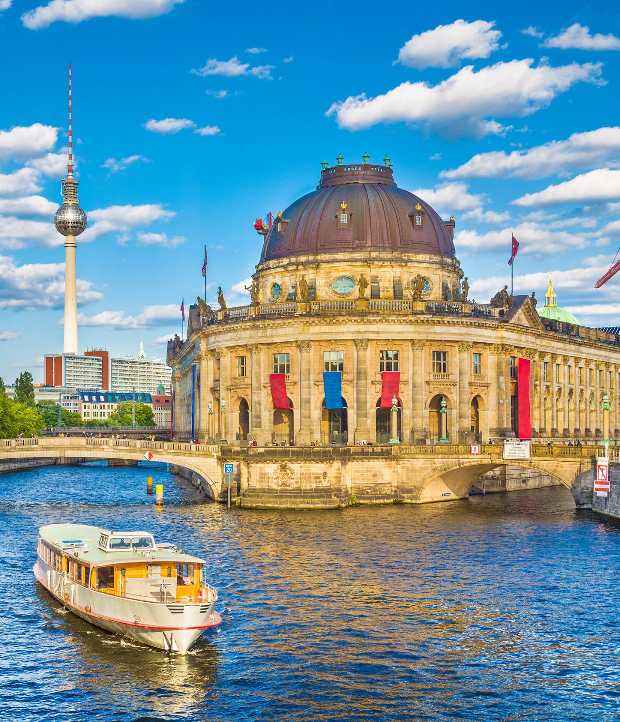 Cidade de Berlim, com uma ponte pedonal sobre um canal, um barco turístico, o museu Bode, e a Torre de Televisão no fundo