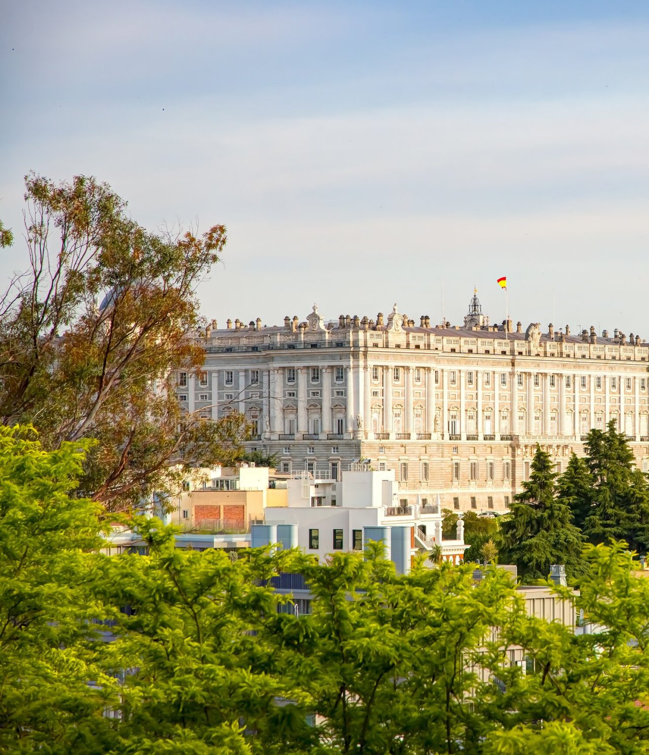 Explore Hotéis em Madrid com vista para o Palácio Real iluminado entre árvores verdes sob céu azul claro