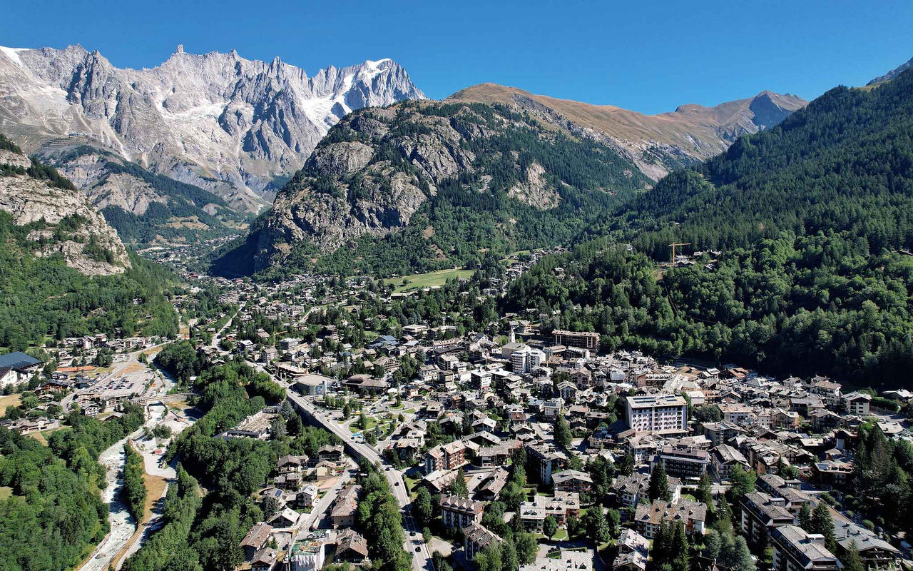 Vista ampla de vila alpina entre montanhas rochosas e florestas densas sob céu azul limpo no vale de Courmayeur