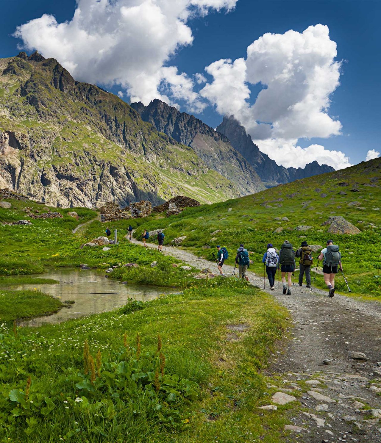 Caminhantes percorrem trilho alpino entre prados verdes e montanhas rochosas sob nuvens densas brancas