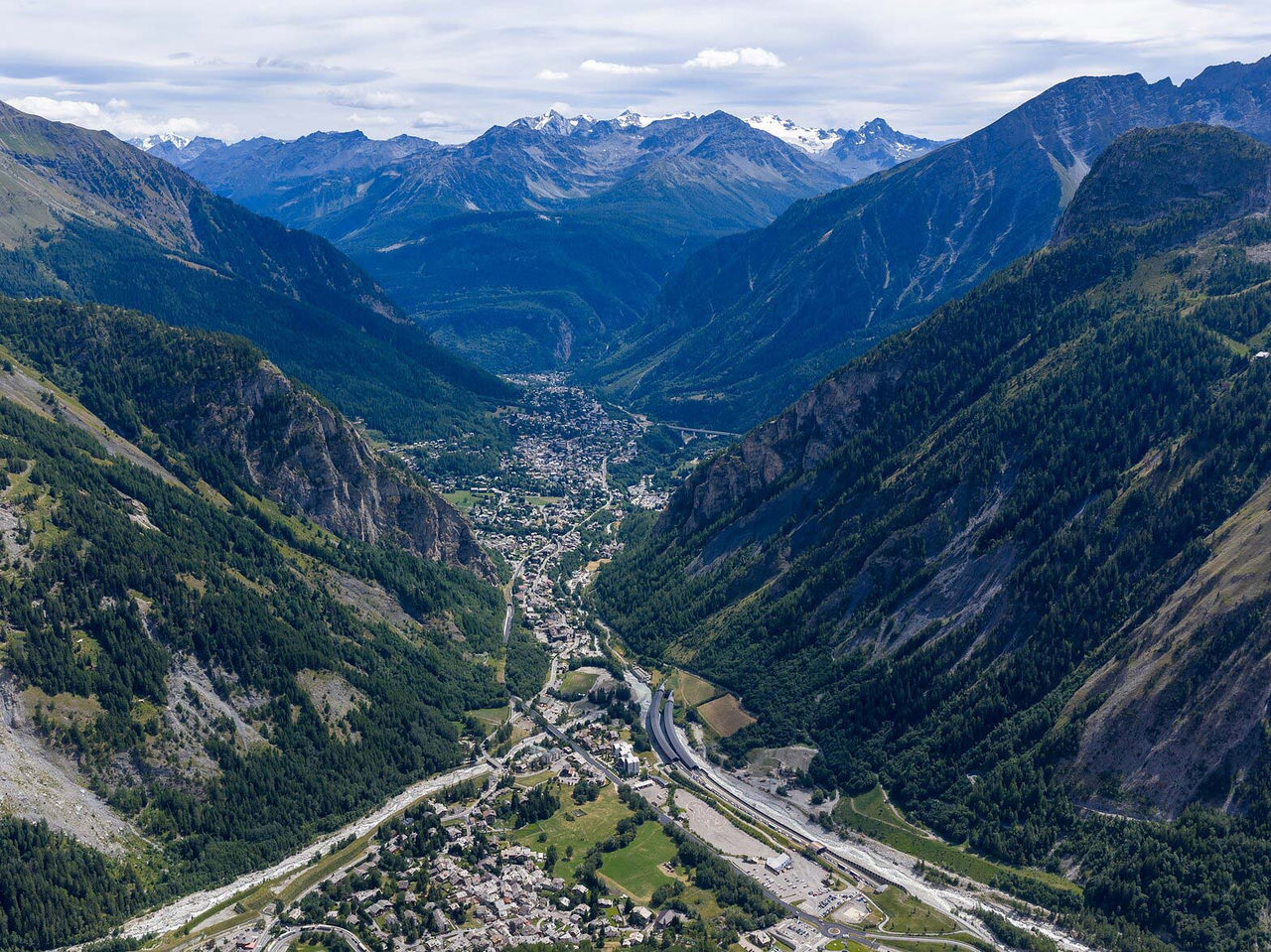 Vista aérea de vale alpino em Courmayeur com montanhas arborizadas, estrada sinuosa e vila ao fundo