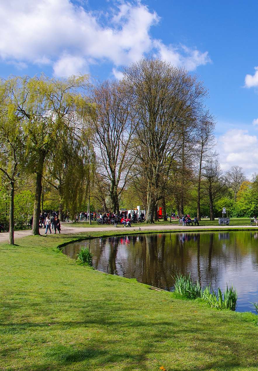 Vista panorâmica do Vondelpark num dia de sol, com um lago cercado por relva verde, árvores e pessoas a relaxarem