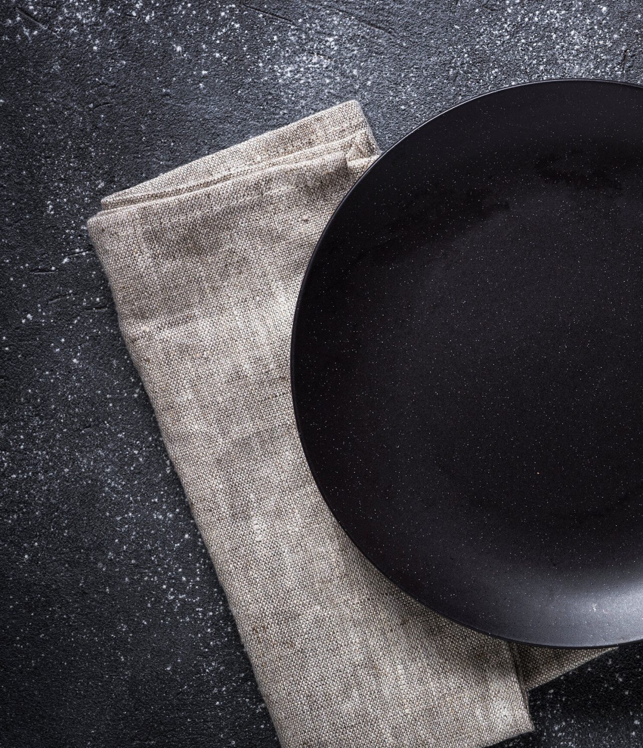 Prato gourmet de jantar, de cor preto mate, colocado sobre um guardanapo cinza claro, em fundo escuro