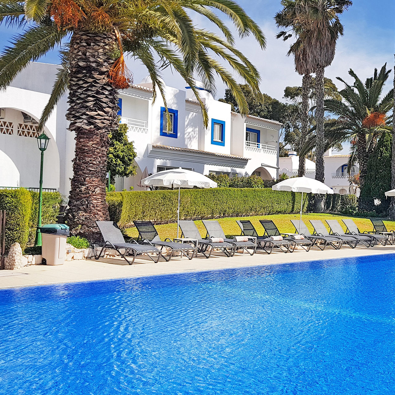 Piscina exterior no hotel no Carvoeiro, com espreguiçadeiras, chapéus de sol, palmeiras e villas atrás