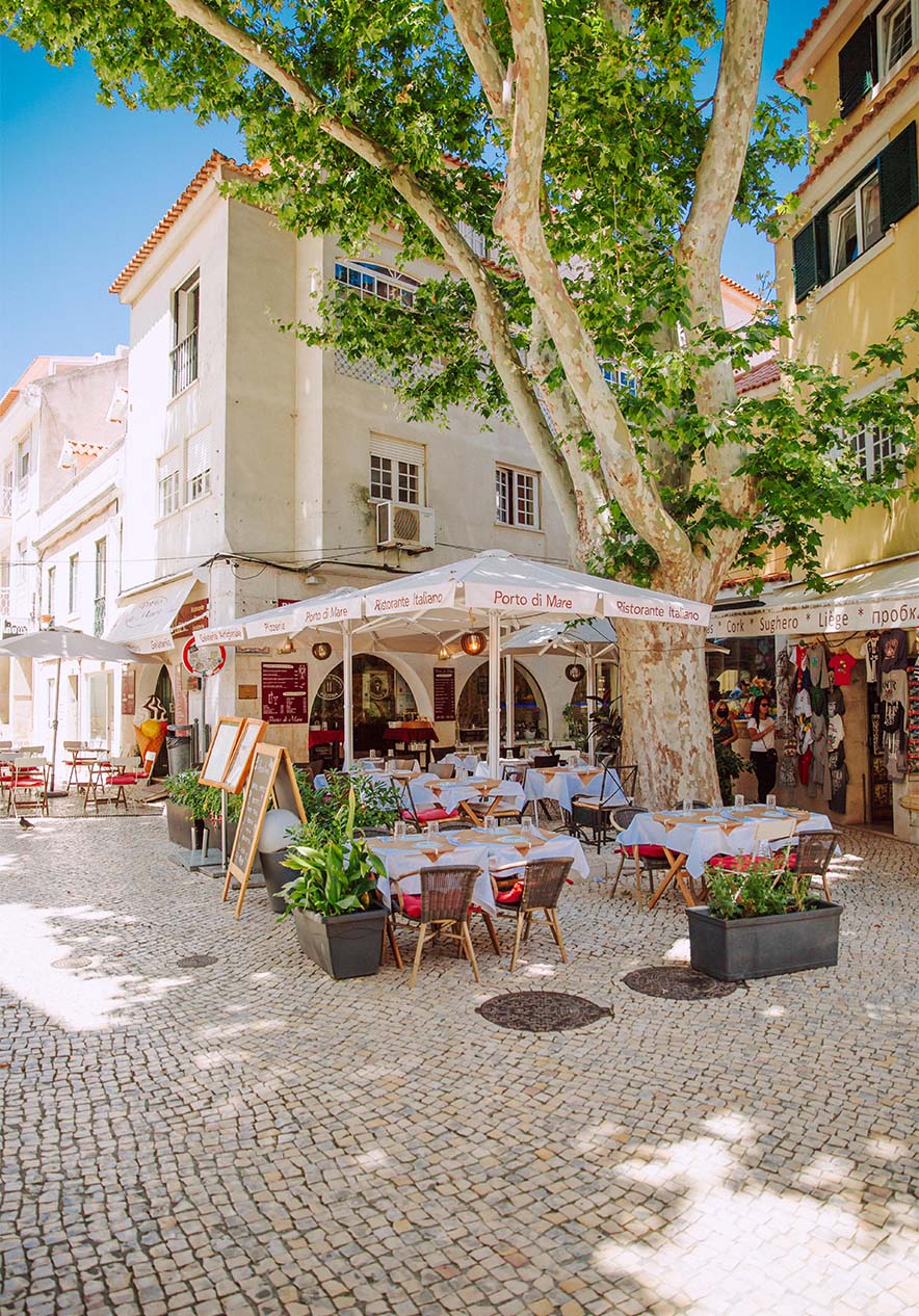 Restaurante ao ar livre no largo do Camões, em Cascais, com mesas e cadeiras sob uma grande árvore, em um dia ensolarado.