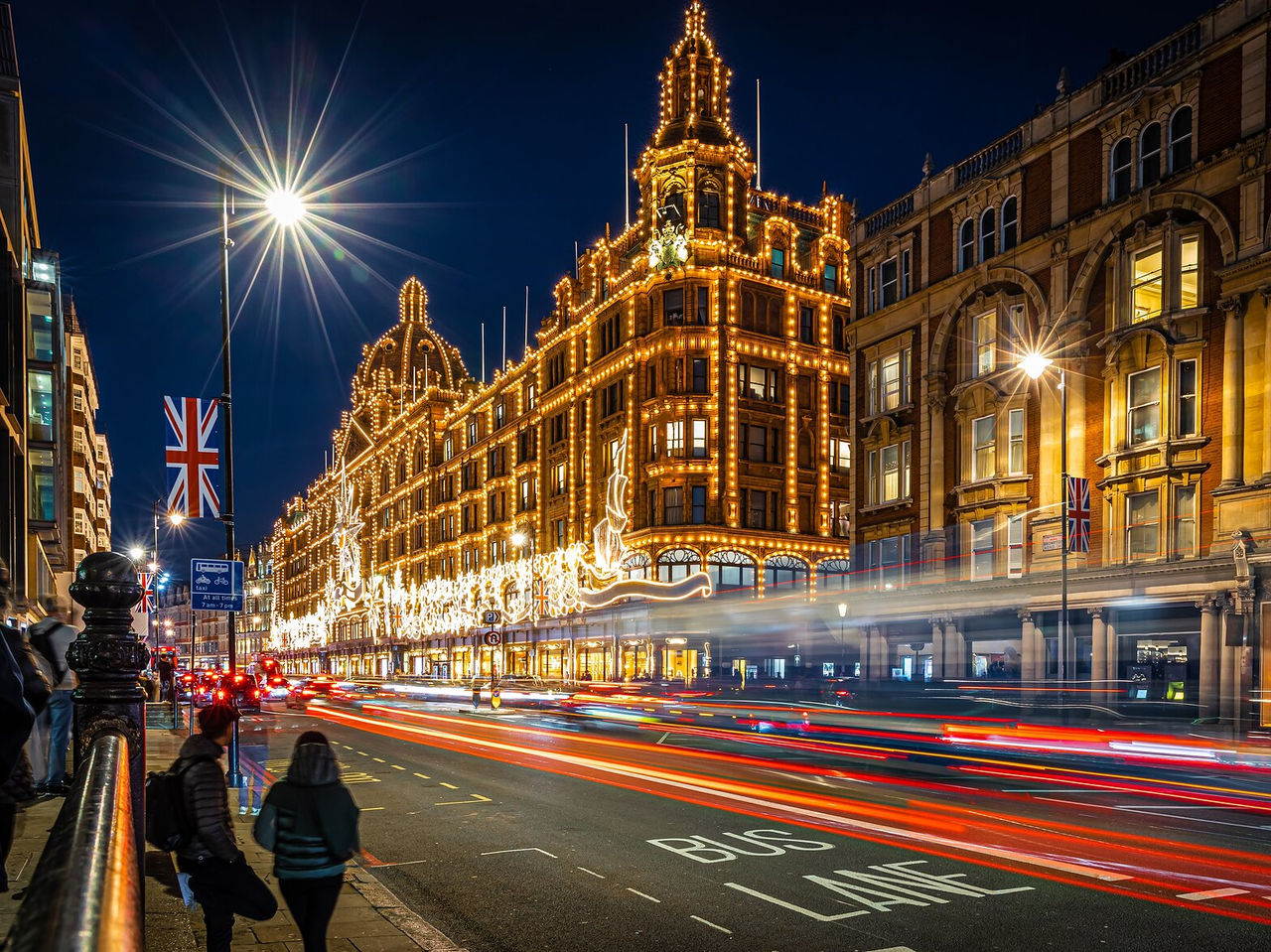 Agitação da cidade Londres, com edifícios iluminados com luzes, trânsito, pessoas a passear e bandeiras de Inglaterra