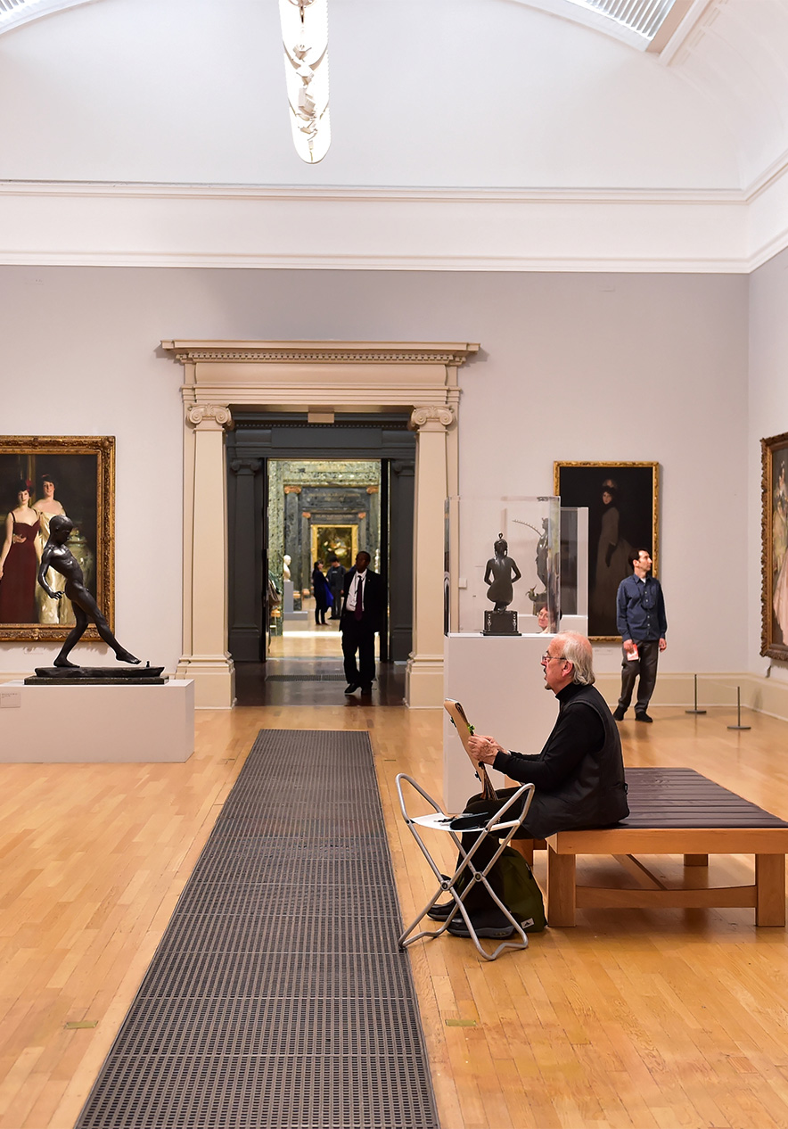 Sala de um museum com diversos quadros e esculturas, pessoas a caminhar, chão de madeira e paredes brancas