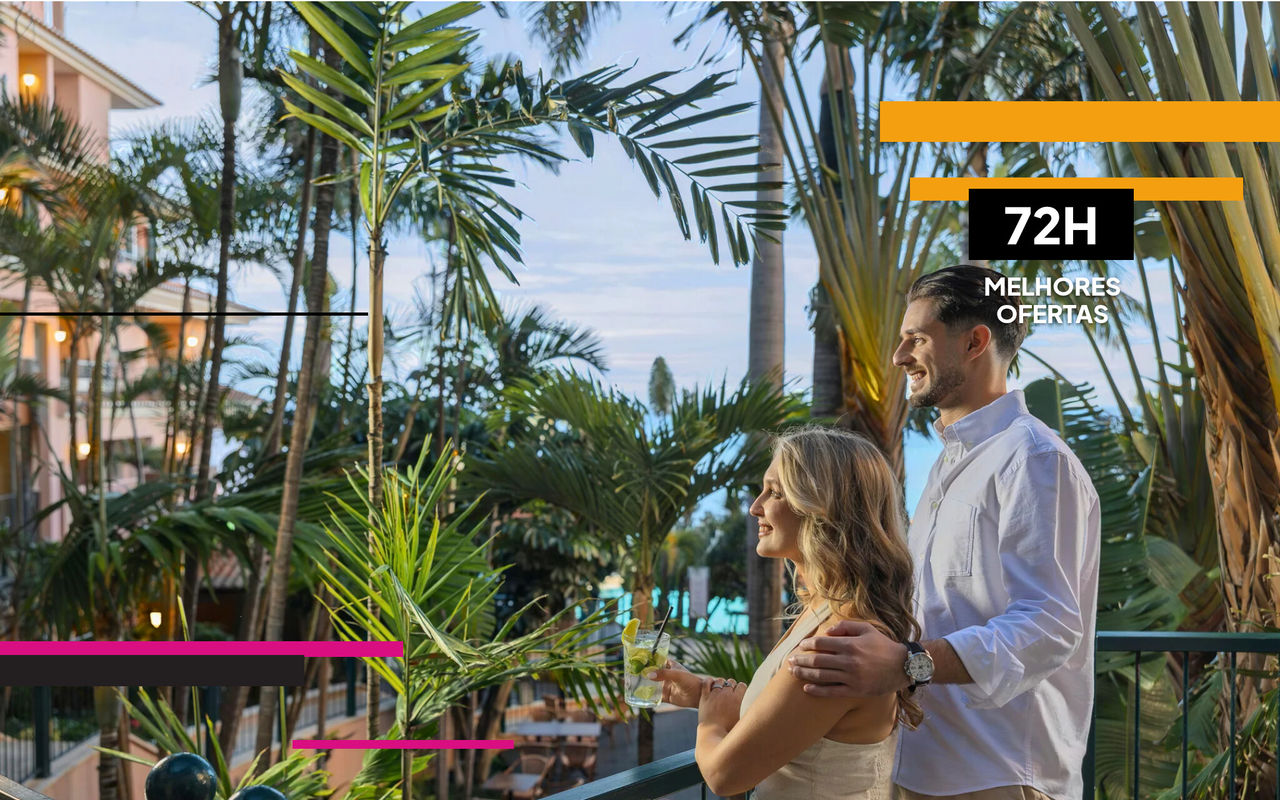 Casal junto a varanda tropical com bebidas ao entardecer entre palmeiras e vista para a piscina do hotel