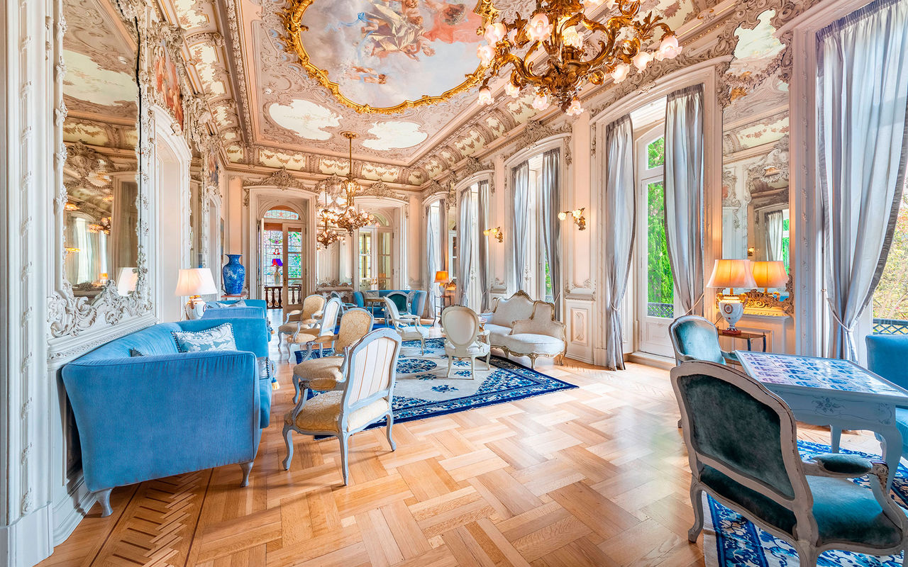 Sala elegante com decoração clássica, lustres dourados, teto pintado e mobiliário azul e branco