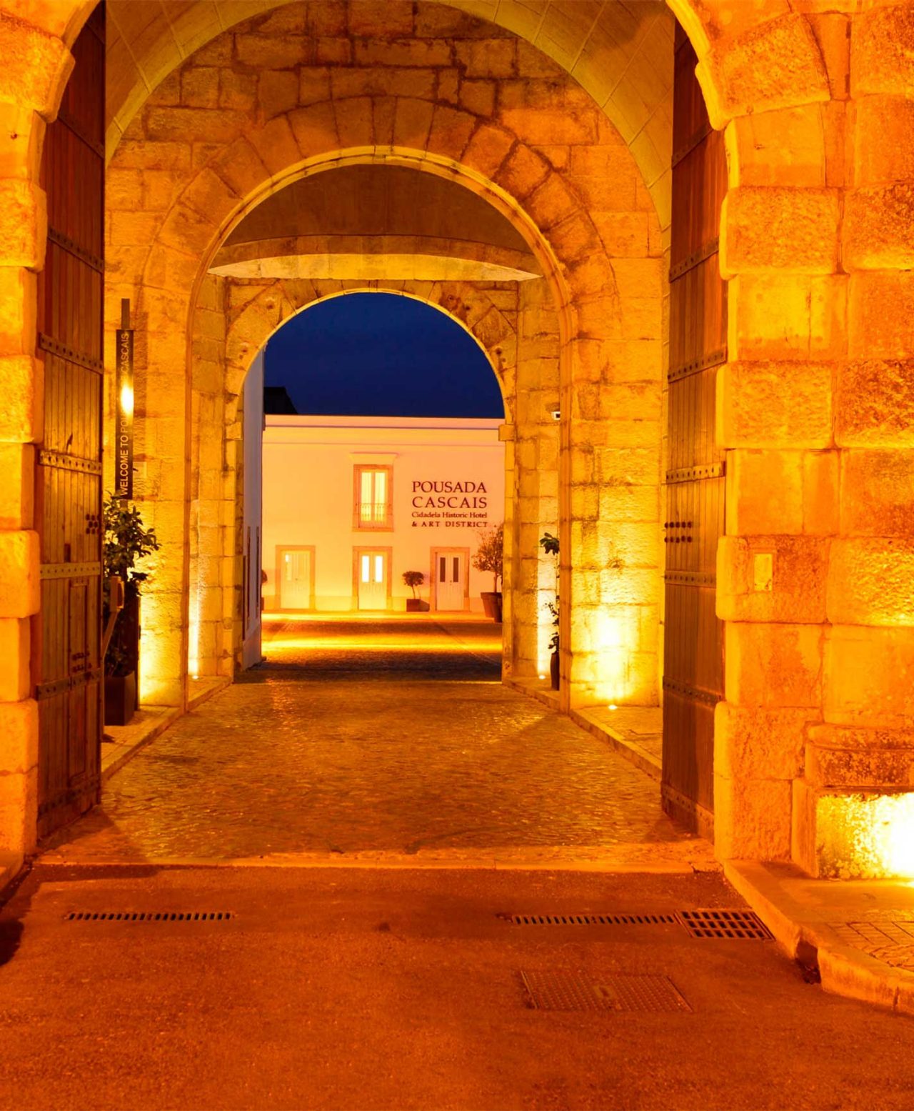 Arco de pedra iluminado à noite com vista para o edifício da Pousada de Cascais
