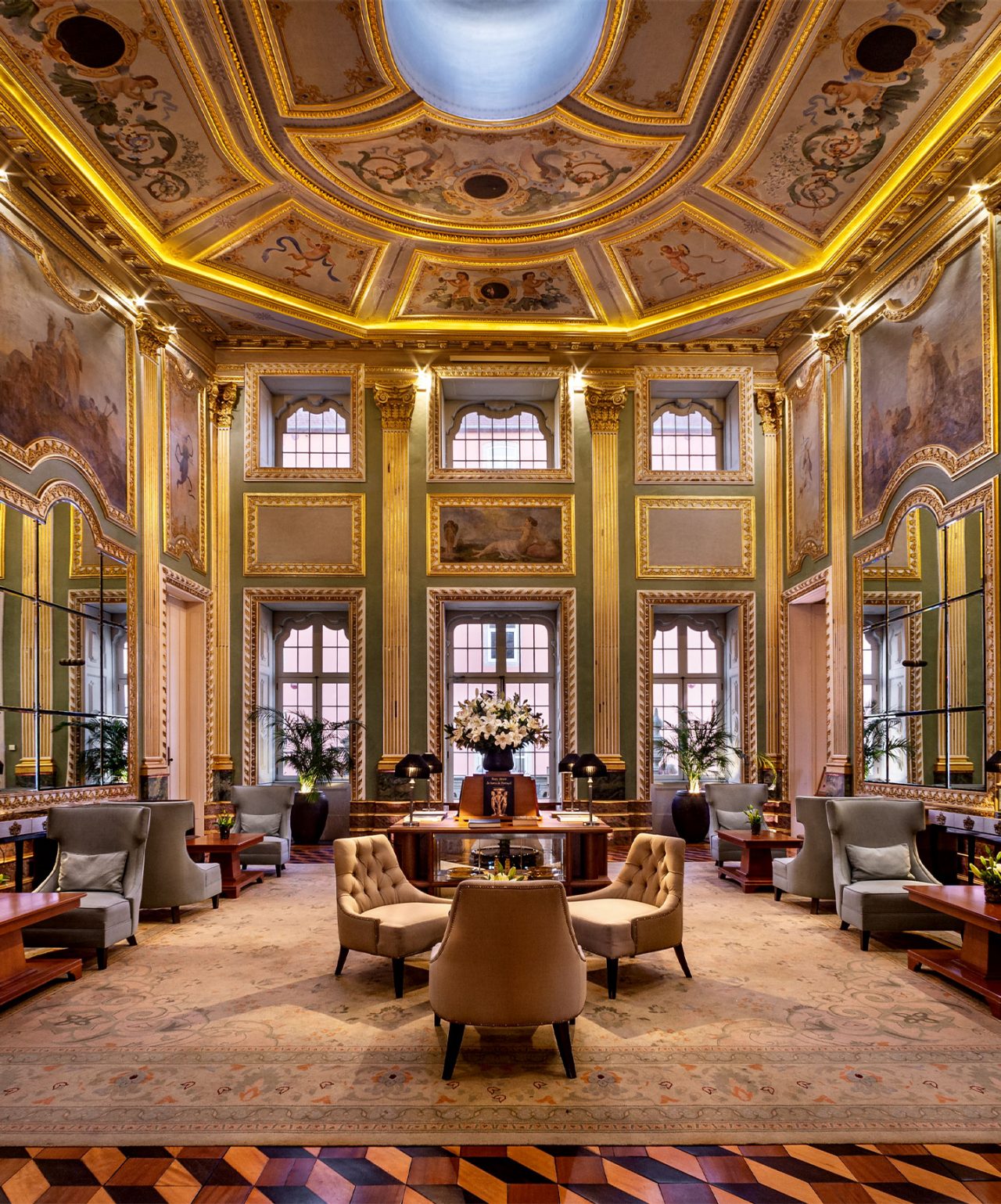 Sala elegante com teto ornamentado e pinturas clássicas, sofás e poltronas dispostos em ambiente luxuoso