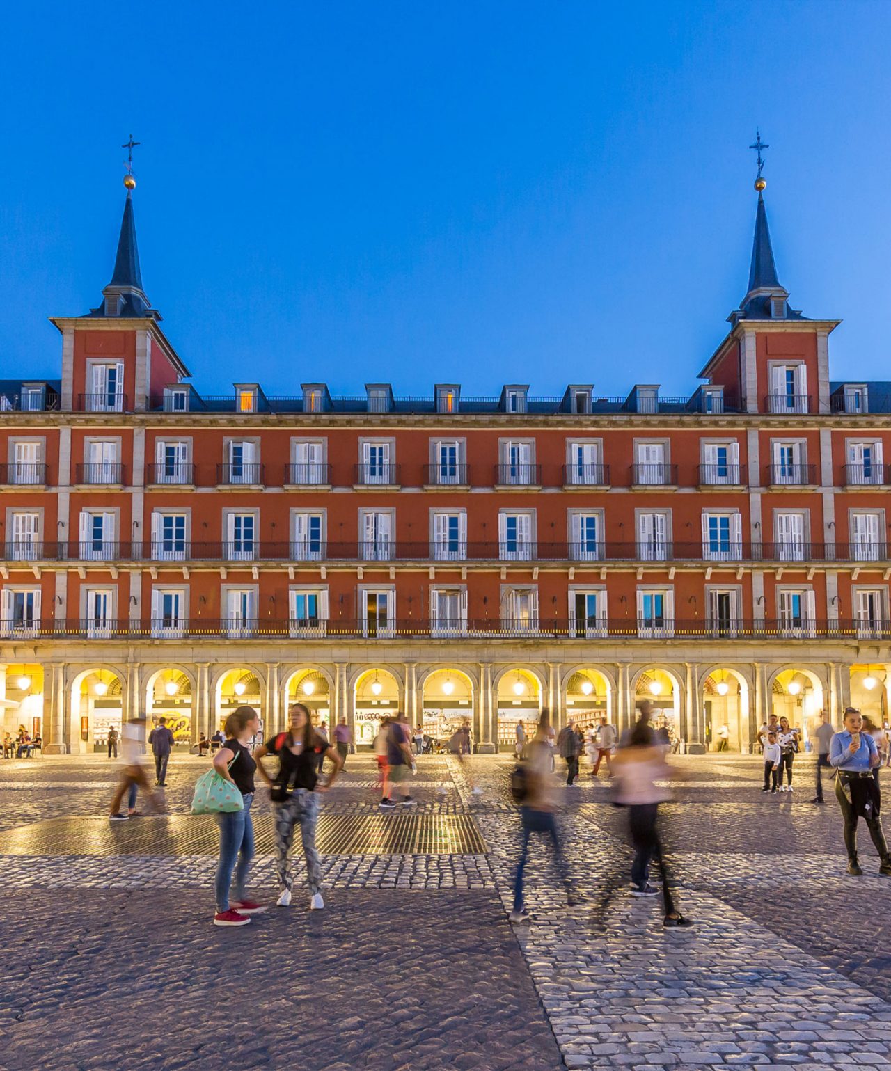 Fachada iluminada da Plaza Mayor em Madrid ao entardecer, com pessoas caminhando na praça