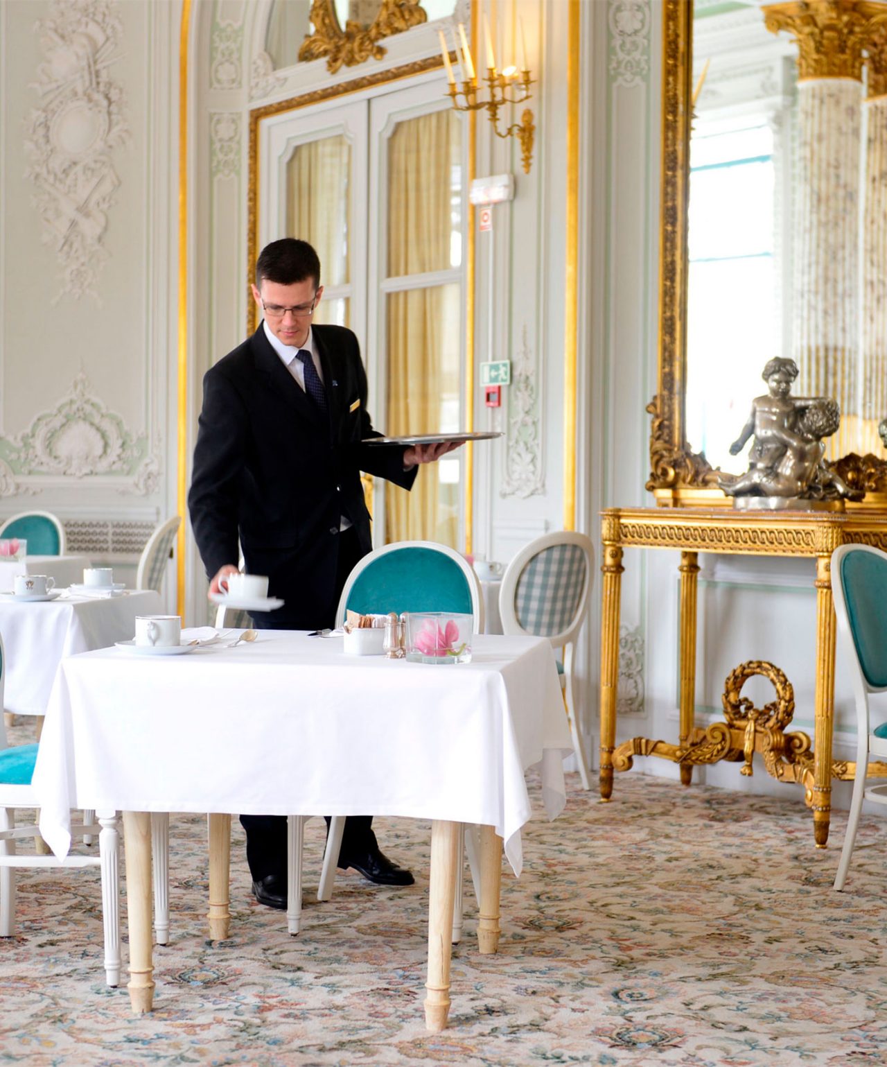 Empregado de mesa a servir chá numa sala elegante com decoração clássica e detalhes dourados