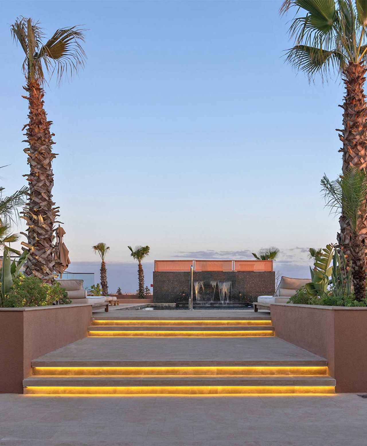 Escadas iluminadas com luz amarela, palmeiras e fonte decorativa ao fundo sob céu azul