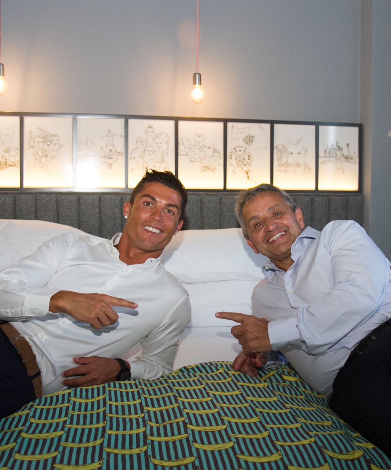 Dois homens sentados numa cama com colcha colorida, apontando um para o outro, com quadros e lâmpadas ao fundo