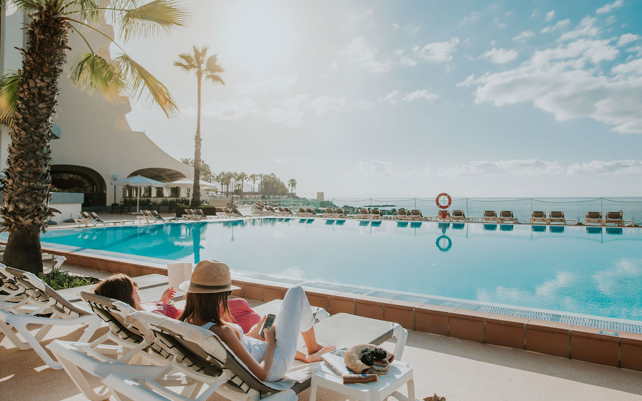 Mulheres relaxando em espreguiçadeiras junto à piscina de hotel, com palmeiras e vista para o mar sob céu azul