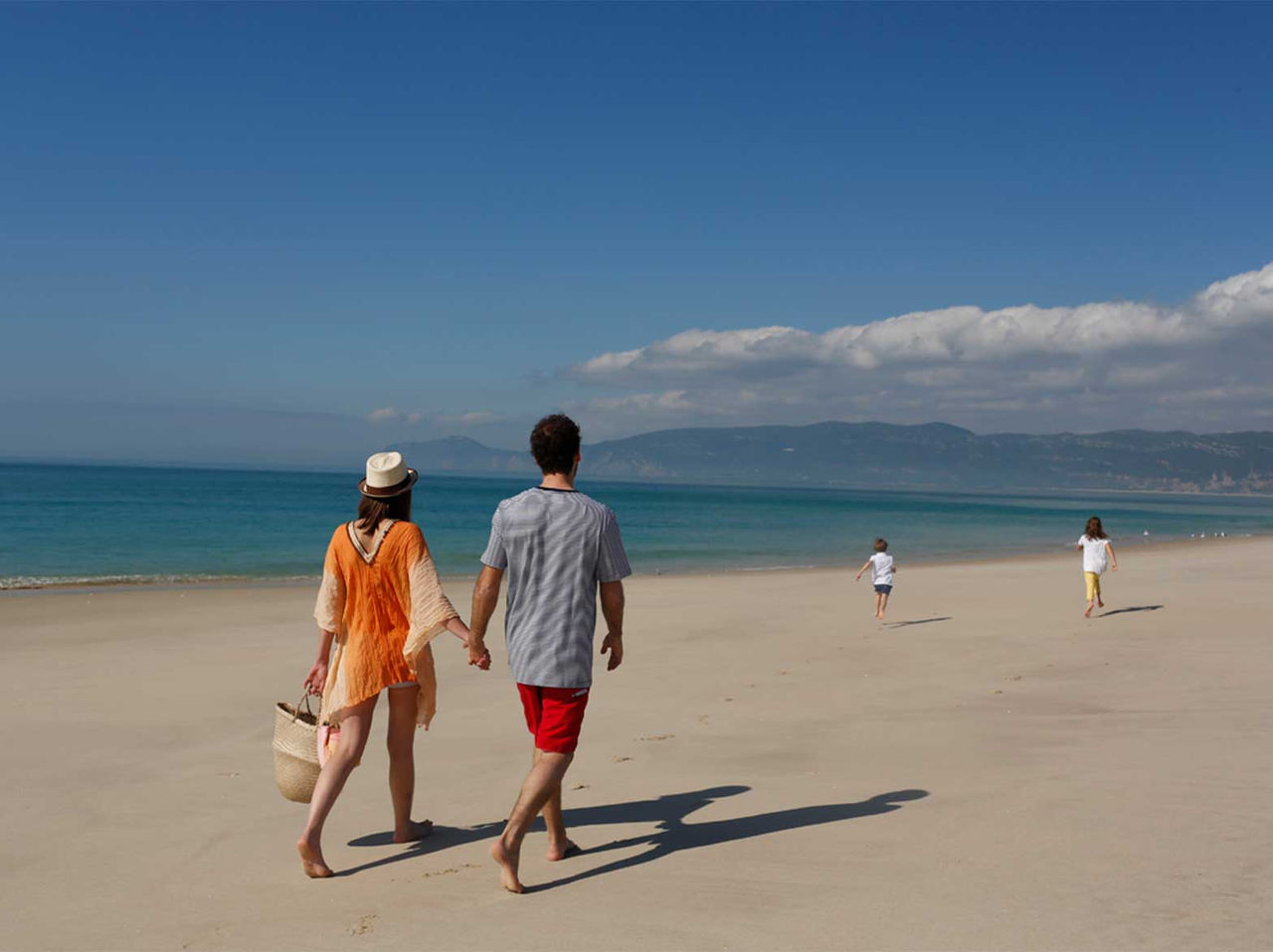 Casal a caminhar na areia junto ao mar, com duas crianças à frente na praia de Troia sob céu azul