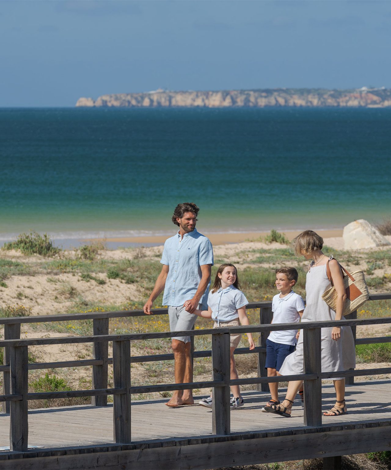 Família a passear num passadiço de madeira junto à praia com o mar azul ao fundo