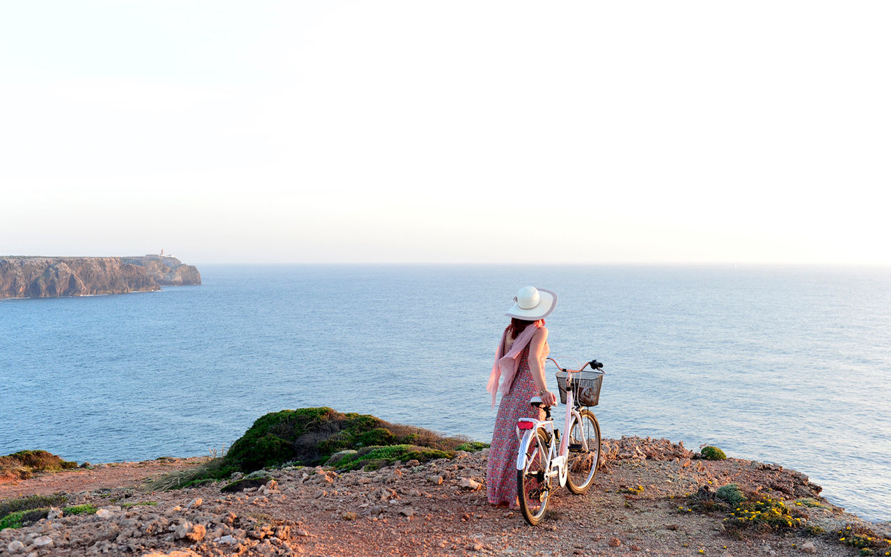 Mulher passeia com bicicleta na mão, junto a arribas na costa portuguesa, disponibilizada pelas Pousadas de Portugal