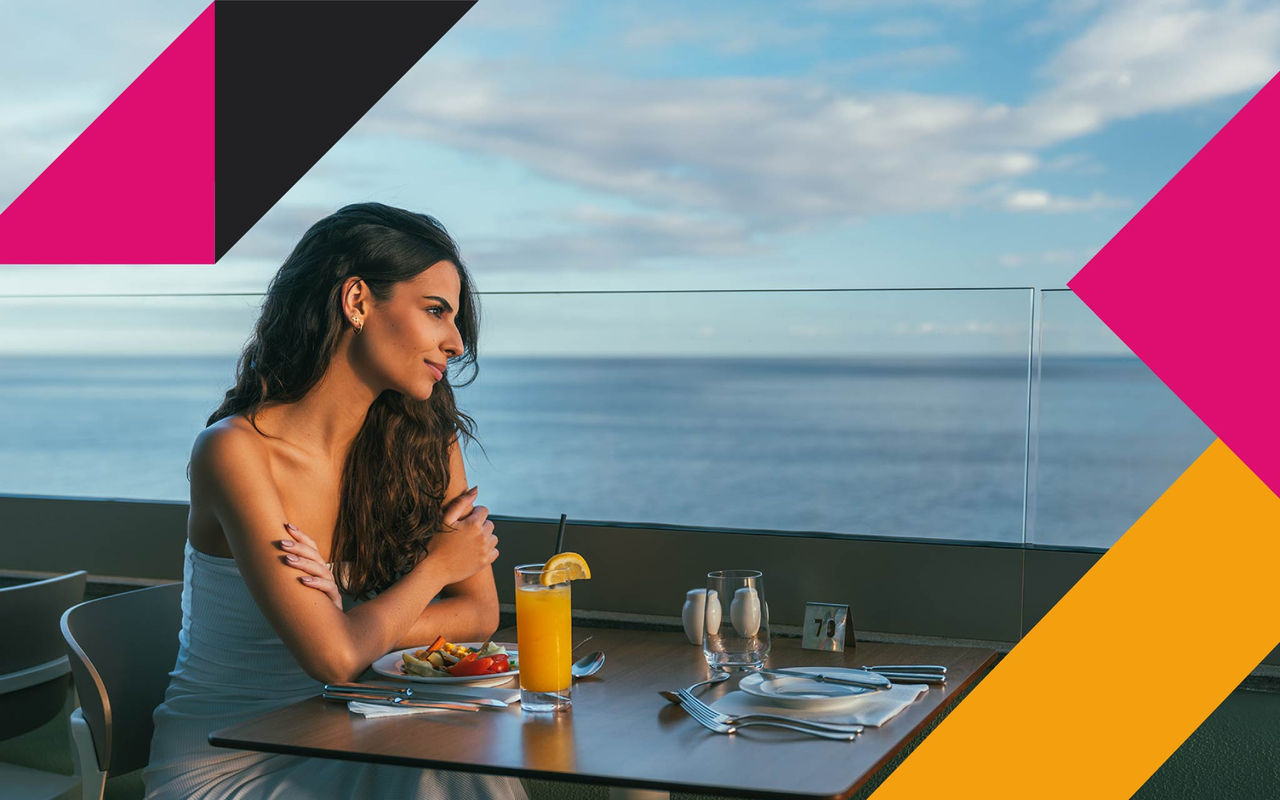 Mulher sentada à mesa com refeição e sumo de laranja, vista para o mar ao fundo sob céu parcialmente nublado