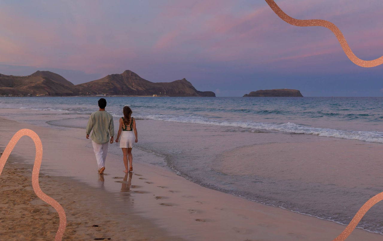 Casal a caminhar de mãos dadas na praia ao entardecer com montanhas e mar calmo ao fundo sob um céu rosado