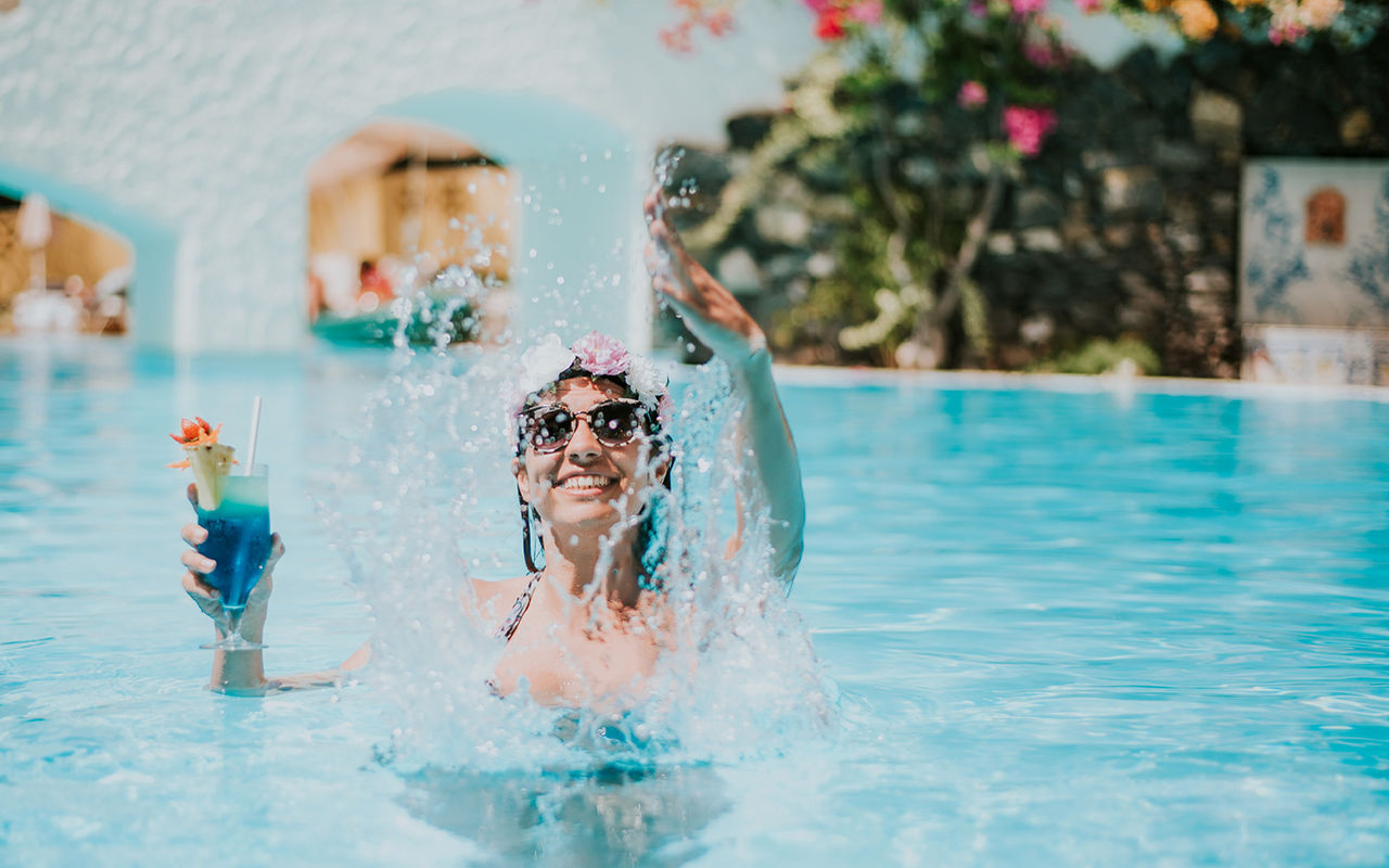 Pessoa sorridente numa piscina azul segurando um cocktail tropical enquanto salpica água num dia ensolarado