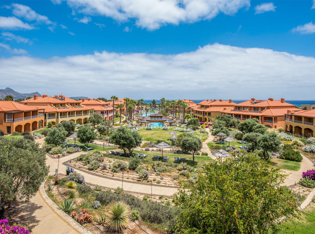 Vista ampla de resort com jardins, piscinas centrais, palmeiras e edifícios de telhados laranja junto ao mar