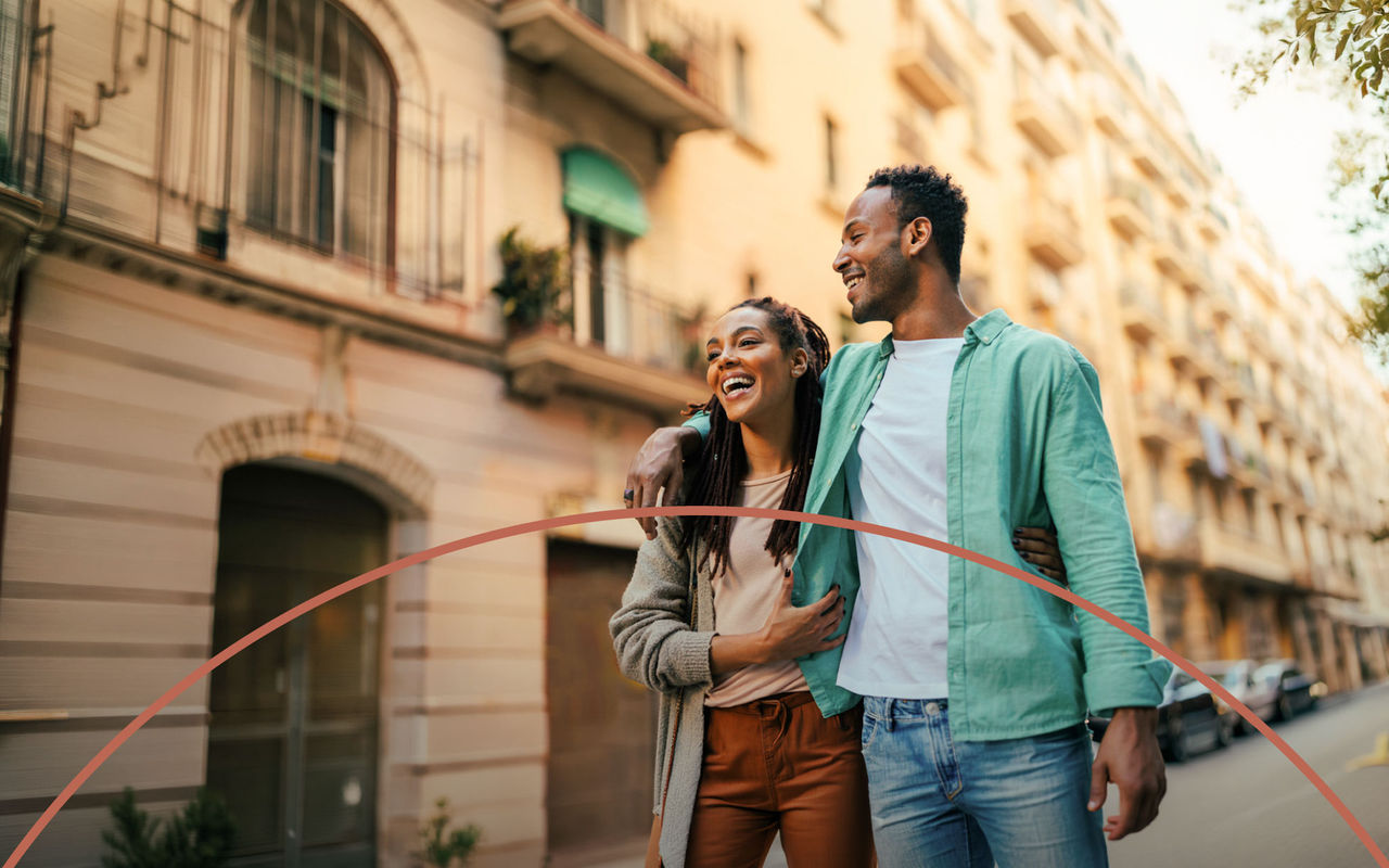 Casal sorridente a caminhar abraçado por uma rua urbana iluminada, com prédios residenciais ao fundo
