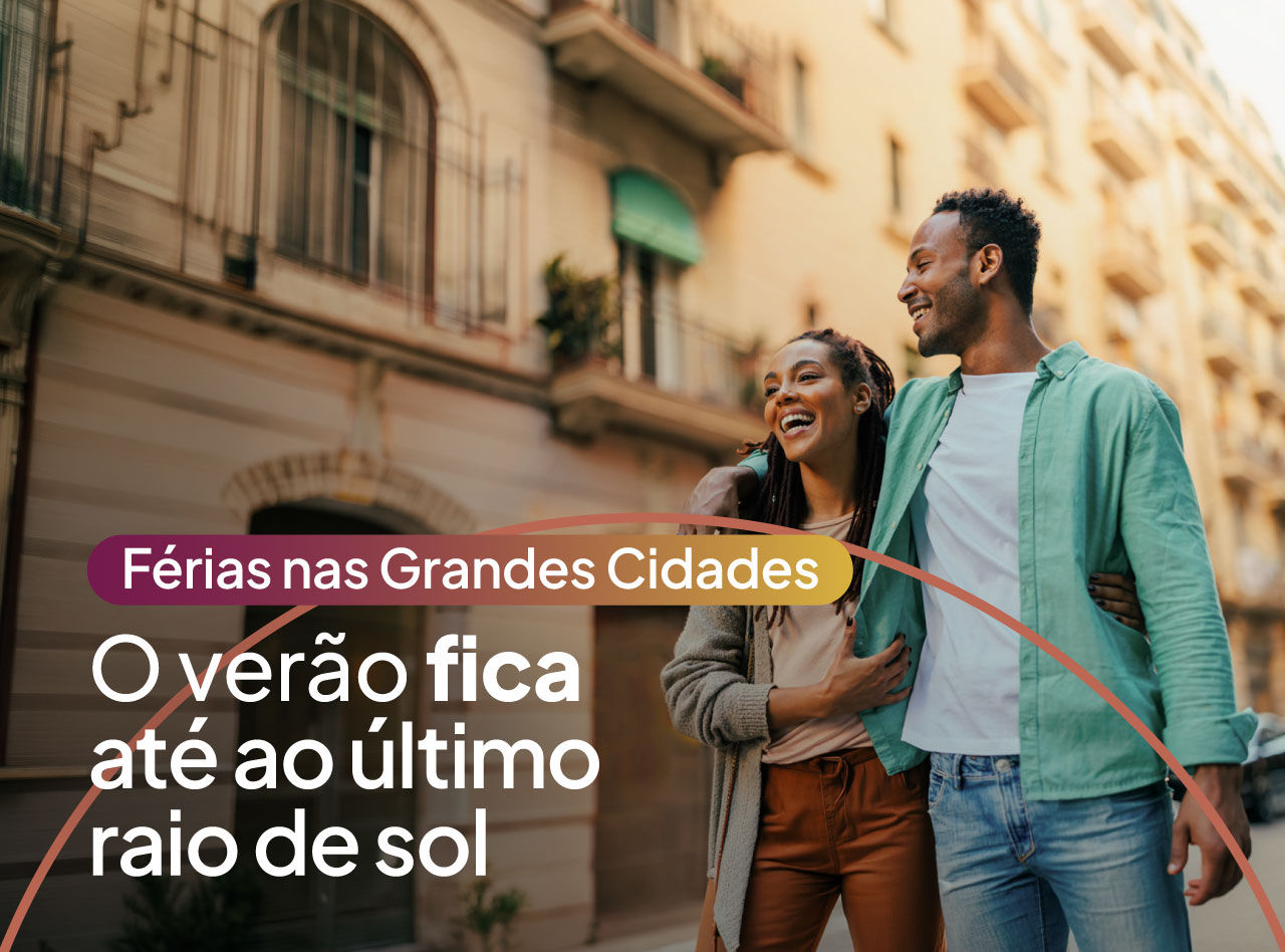Casal sorridente caminha por rua urbana ao entardecer entre edifícios iluminados pelo sol de verão quente