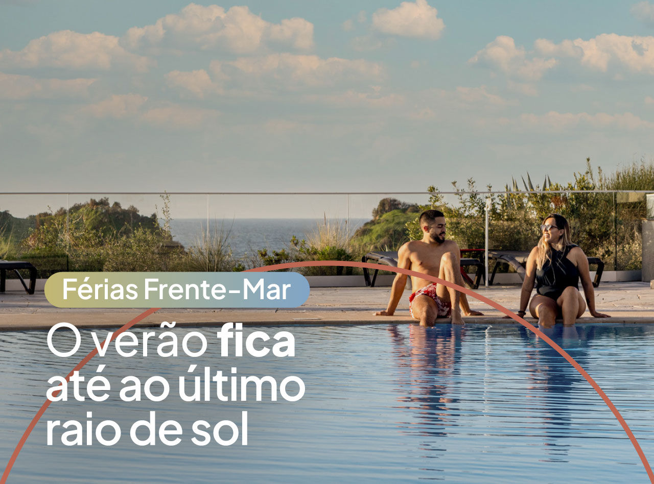 Casal sentado na borda de uma piscina ao ar livre com vista para o mar sob céu azul com nuvens ao entardecer