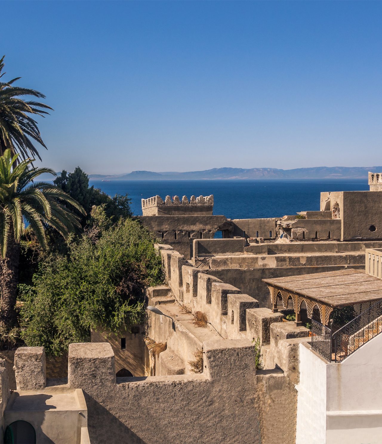 Explore Hotéis em Tânger com vista para muralhas históricas junto ao mar azul e palmeiras sob céu limpo