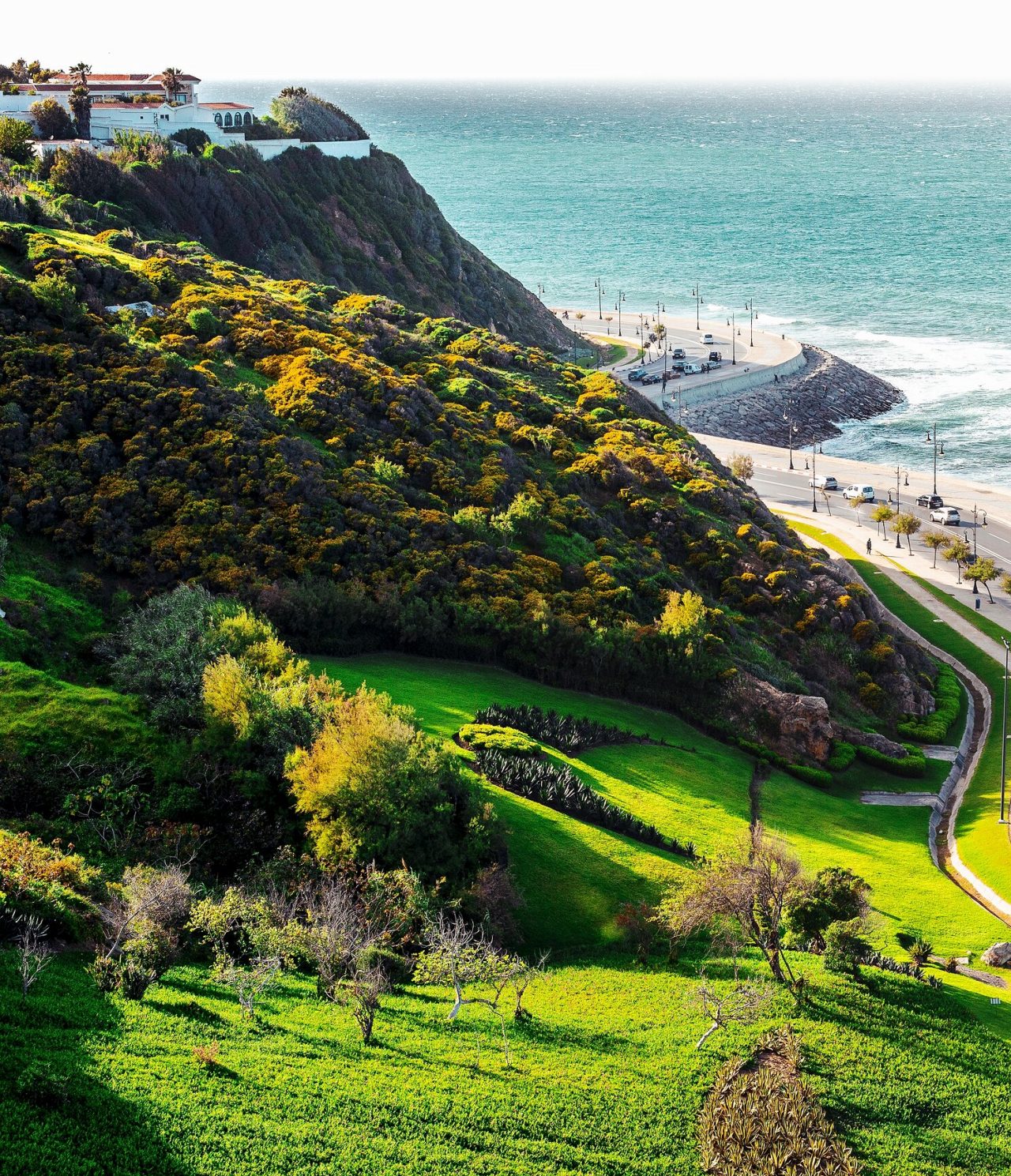 Explore Hotéis em Tânger com vista para falésias verdes junto à costa e estrada costeira banhada pelo mar