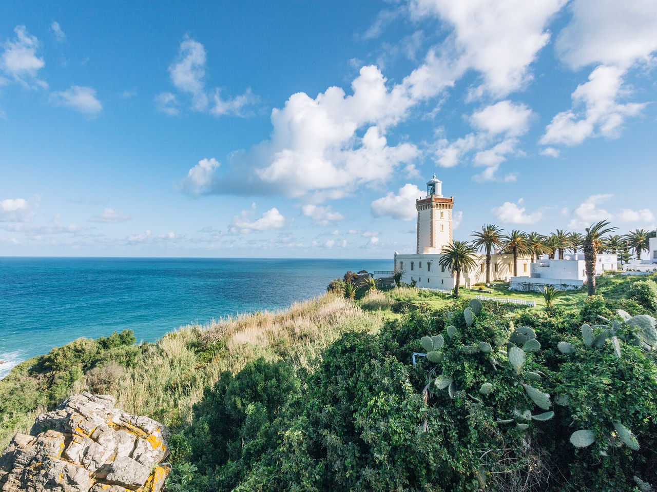 Explore Hotéis em Tânger com vista para farol branco junto ao oceano e vegetação costeira sob céu azul