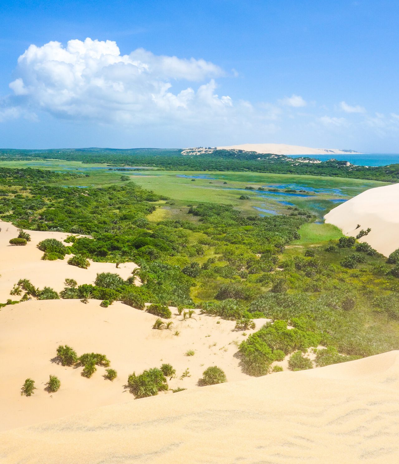 Aerial view of a desert landscape with white sand dunes, a lagoon, and crystal-clear ocean waters in the background